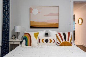 Un dormitorio con cama, almohadas, mesita de noche, lámpara y un cuadro en la pared.