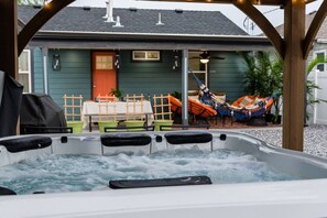 A hot tub with a wooden pergola overhead, a dining table with chairs, and a hammock outside a house.