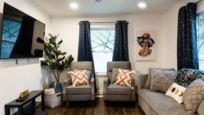 A living room with two armchairs, a sofa, a TV, a large window with curtains, and a plant. - Rise & Grind SLC HIGH-END Style/Gated n Garage-2Br (South Salt Lake)
