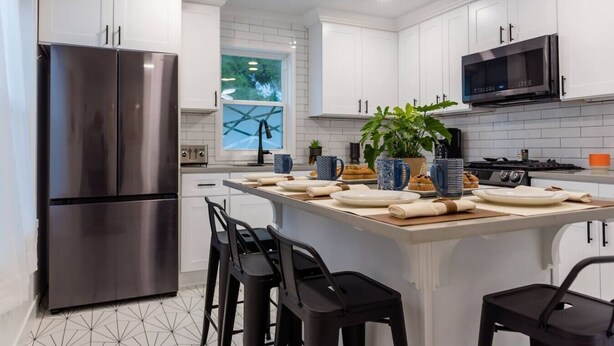 A modern kitchen with white cabinets, stainless steel appliances, a central island with bar stools, and a large window. - Rise & Grind SLC HIGH-END Style/Gated n Garage-2Br (South Salt Lake)
