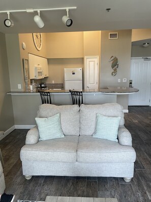 A living room with a grey sofa, two black chairs, and a kitchen area with a fridge, microwave, and a decorative wall piece.