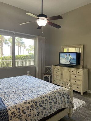 A bedroom with a bed, a ceiling fan, a television on a dresser, and a window with a view of palm trees.