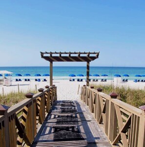 A wooden boardwalk leading to a beach with blue umbrellas and lounge chairs.