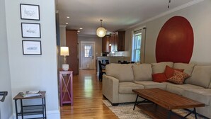 A spacious living room with a wooden floor, a beige sofa, a wooden coffee table, and a red accent wall. - Casa Rosa - 2BR West End Bungalow (Durham)