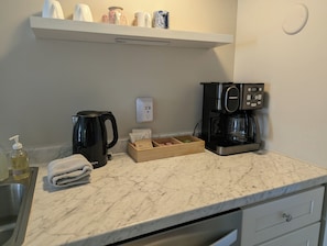 A kitchen with a coffee maker, kettle, and a box of coffee pods. - The Trinity Garden Suite, King Bed Near Duke (Durham)