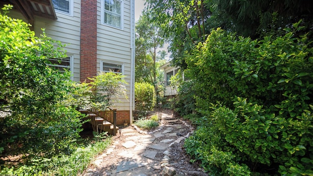 A stone pathway leading to a house with a brick chimney, surrounded by lush greenery.