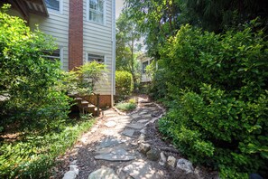A stone pathway leading to a house with a brick chimney, surrounded by lush greenery.