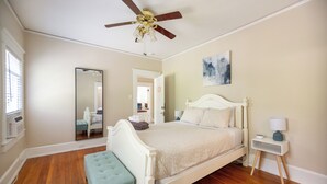 A bedroom with a wooden floor, a white bed, a bench, a nightstand, a ceiling fan, a mirror, and a painting on the wall.