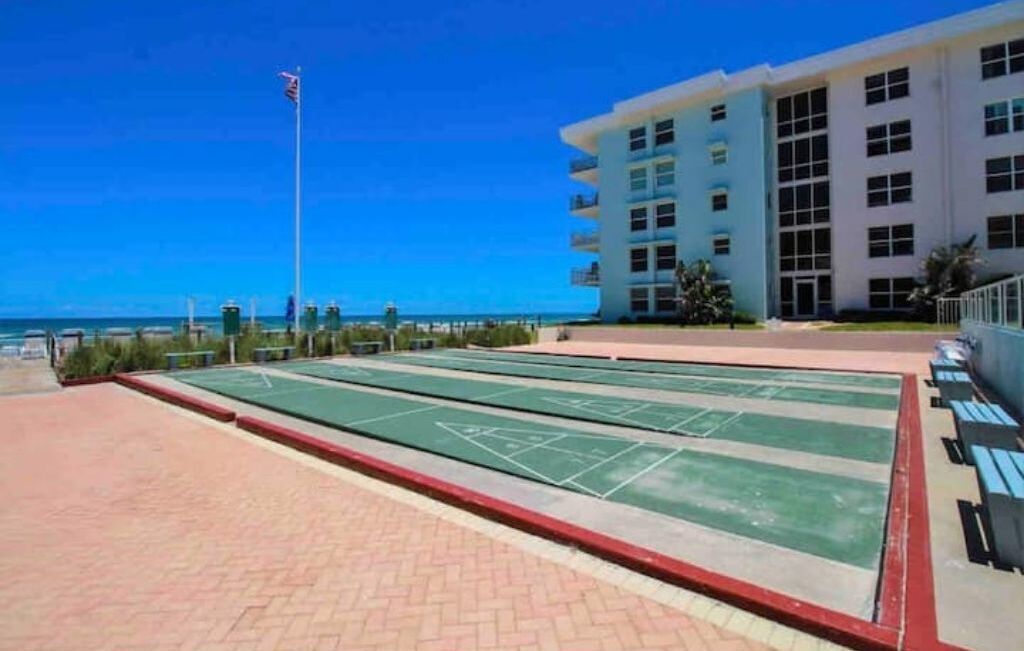 A courtyard with shuffleboard tables, a flagpole, and a building with balconies.