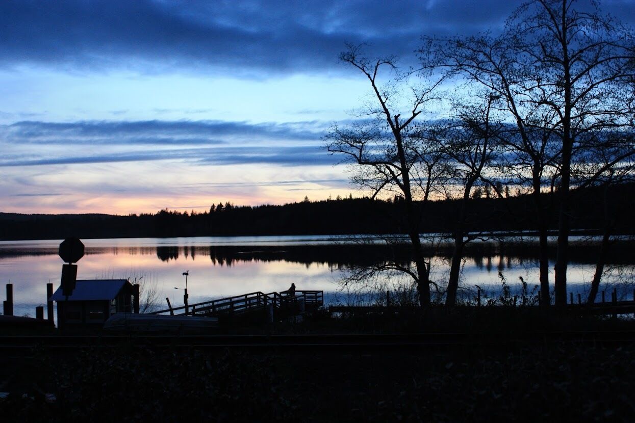 Une scène au bord d’un lac, avec un quai, une petite cabane et des arbres sans feuilles, le tout se détachant sur un ciel crépusculaire.
