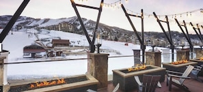 A snow-covered outdoor area with a fire pit and seating, overlooking a ski resort.