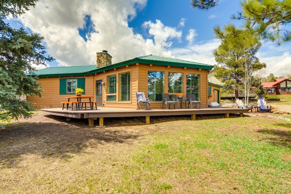 Una cabaña de madera con techo verde, ventanas amplias y una terraza con mobiliario exterior.