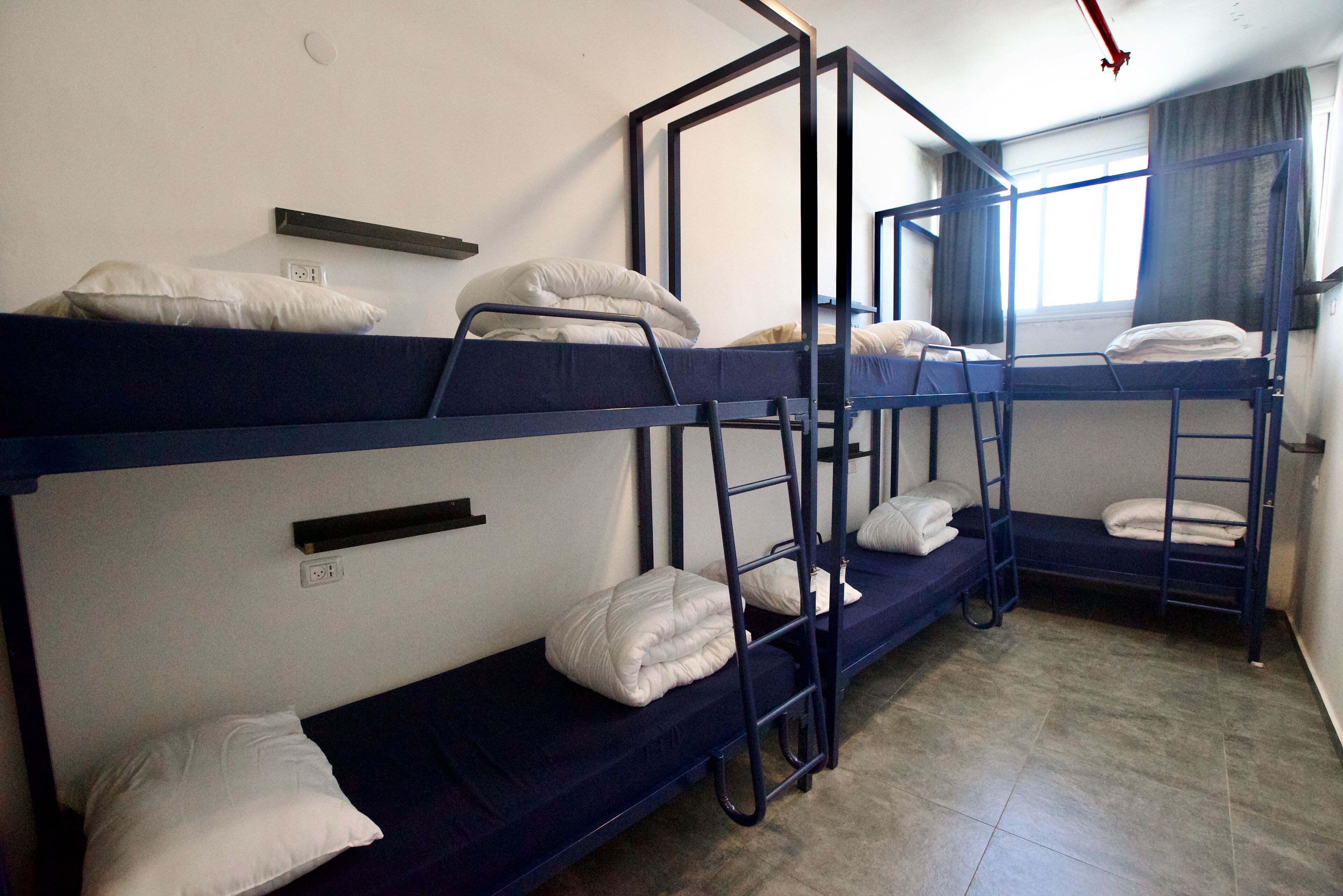 A dormitory room with bunk beds, a shelf above the beds, and a window with curtains.