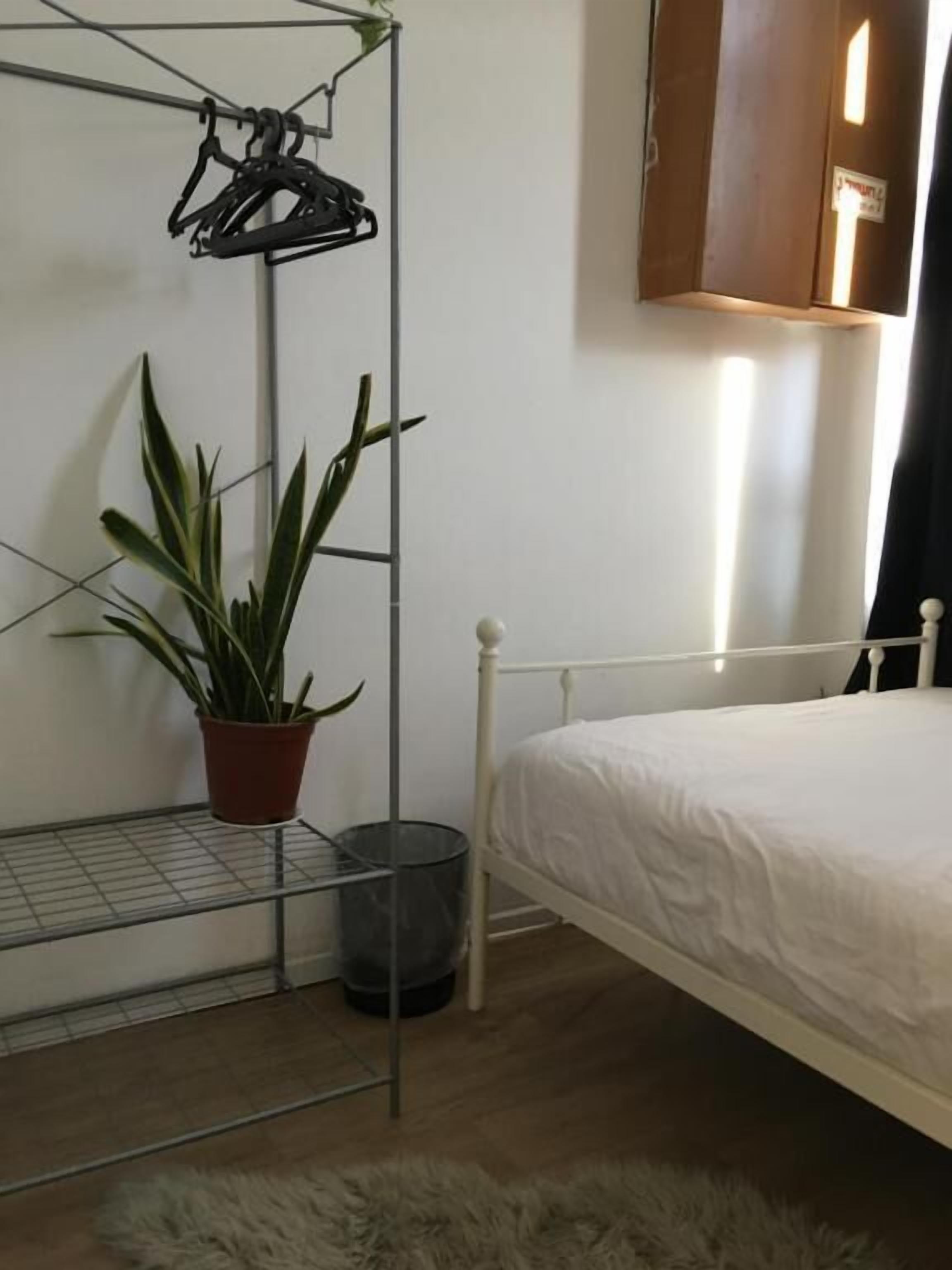 Un dormitorio con una cama blanca, un tendedero de metal, una planta en maceta y un pequeño cubo de basura.