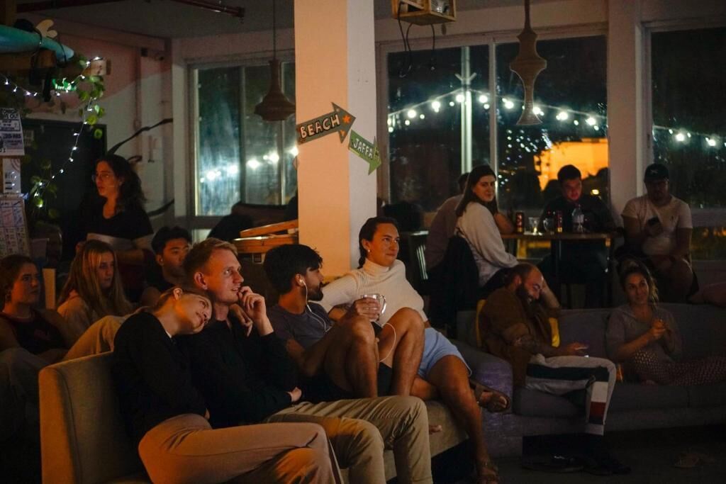 A group of people sitting on a couch in a room with a large window, a sign indicating 'BEACH' and 'JAFRA', and string lights.
