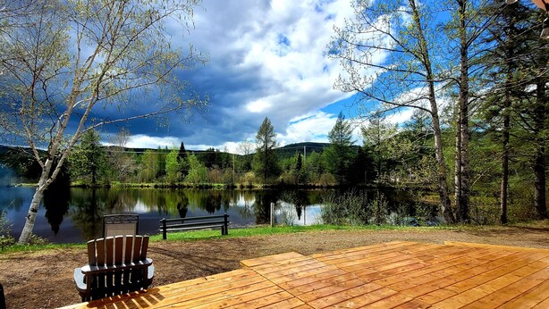 Una terraza de madera con un banco y una mesa, con vista a un lago tranquilo con árboles y una colina distante.