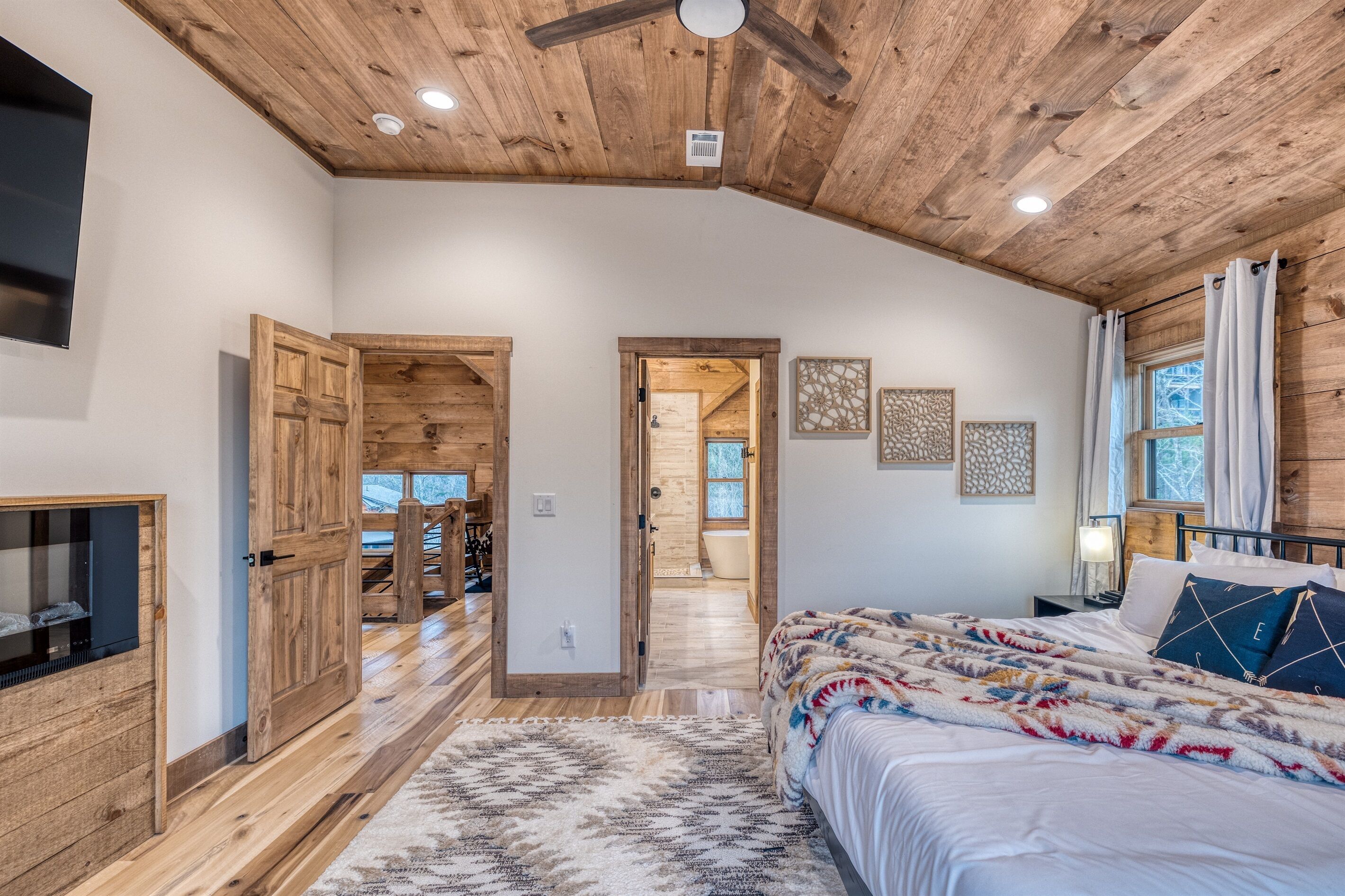 A bedroom with a wooden ceiling, a bed with patterned bedding, a television, and a bathroom visible through an open door.