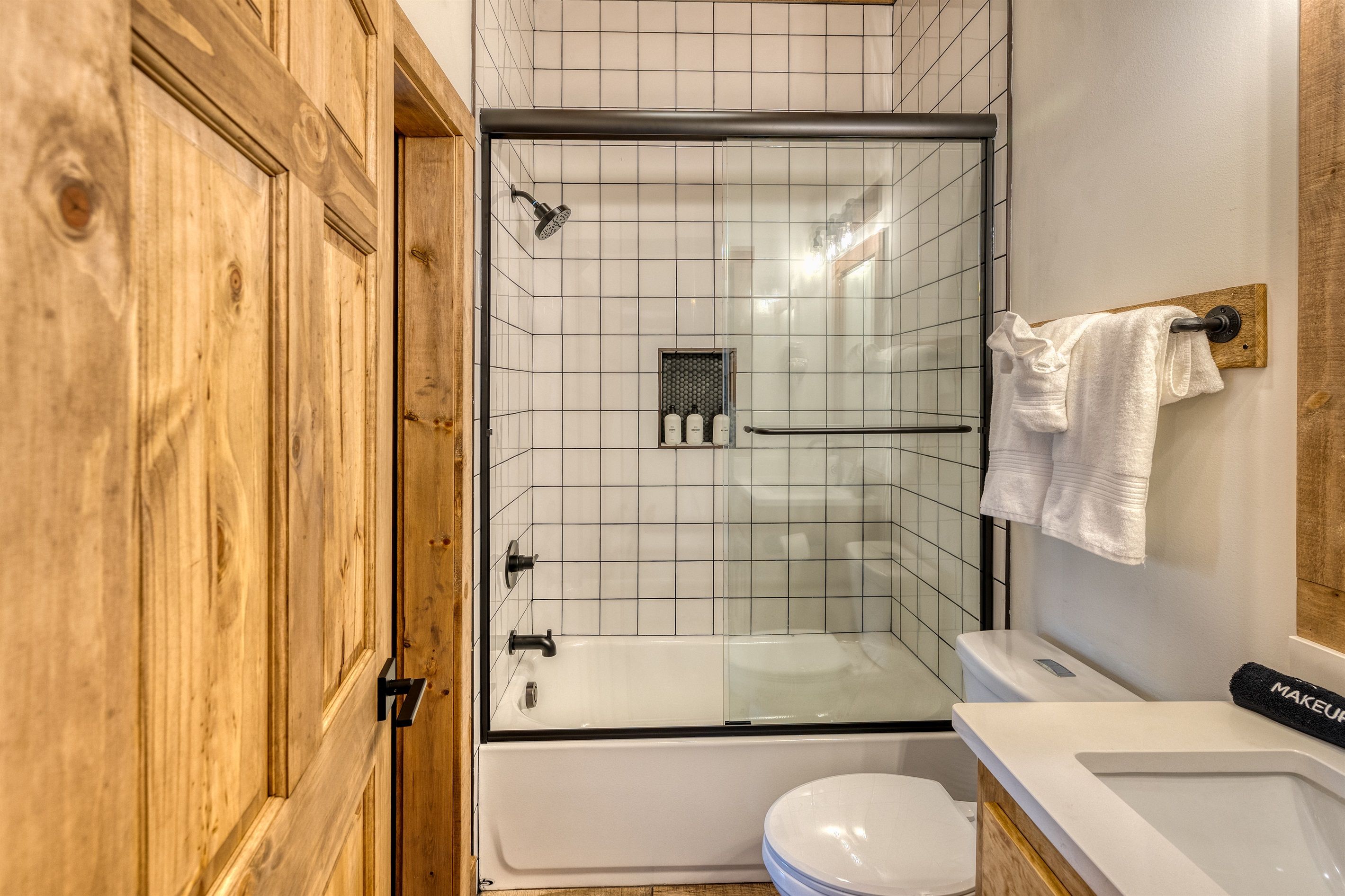 A bathroom with a glass-enclosed shower, a wooden door, a white sink, and a toilet.