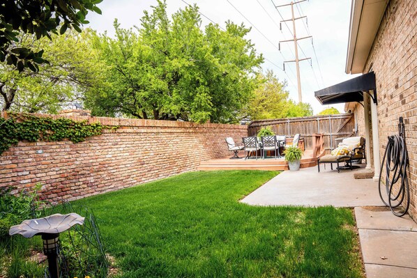 A backyard with a brick wall, a grassy lawn, a patio with outdoor furniture, and a hose coiled against the wall.