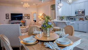 A dining area with a round table set for four, a chandelier, and a kitchen in the background.