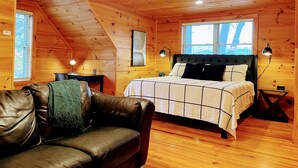 A cozy cabin room with a bed, a leather couch, a ceiling fan, and wooden walls. - 6BR Log Cabin | Walk to Downtown | Mountain Views (West Jefferson)