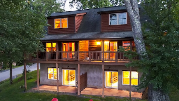 A two-story log cabin with a balcony, multiple windows, and a fire pit area with red chairs. - 6BR Log Cabin | Walk to Downtown | Mountain Views (West Jefferson)