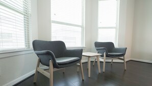 Une pièce avec deux fauteuils gris et une petite table, de grandes fenêtres équipées de stores et un parquet en bois foncé.