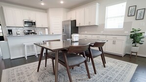 Une cuisine moderne avec des meubles blancs, des appareils en acier inoxydable et un coin repas comprenant une table en verre et des chaises en bois.