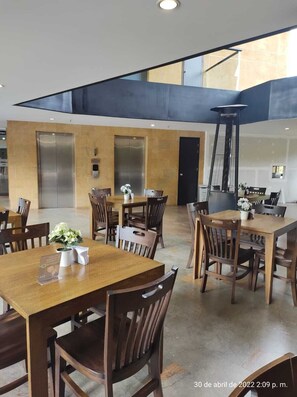 A modern dining area with wooden tables and chairs, two elevators, and a large window providing natural light.
