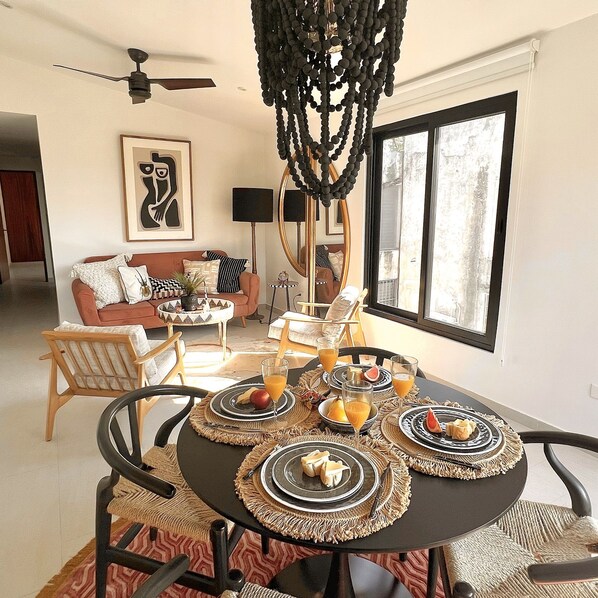 A dining area with a round table set for four, surrounded by chairs. There is a sofa and a chair in the background, and a large mirror on the wall.
