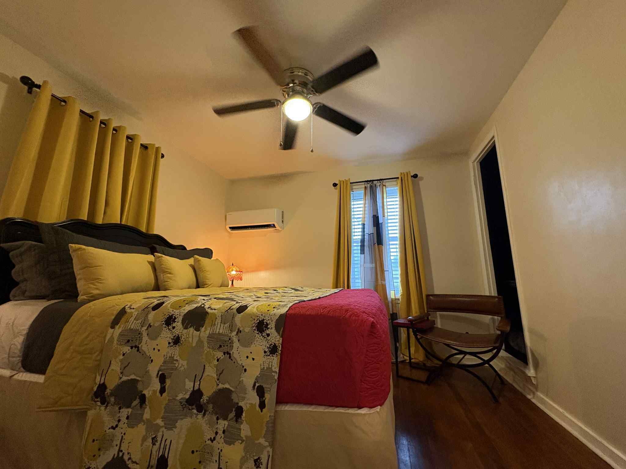 A bedroom with a bed, a ceiling fan, an air conditioning unit, a chair, and a window with curtains.