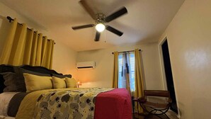 Ein Schlafzimmer mit einem Bett, einem Deckenventilator, einer Klimaanlage, einem Sessel und einem Fenster mit Vorhängen.