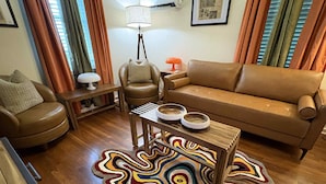 Una sala de estar con un sofá de cuero marrón, un sillón a juego, una mesa de centro de madera con dos cuencos, un tapete colorido y una lámpara de pie.