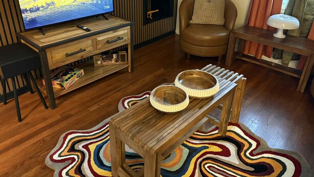 Ein Wohnzimmer mit einem Holz-Fernsehschrank, einem Ledersessel, einem kleinen Tisch mit einer Lampe und einem farbenfrohen Teppich.