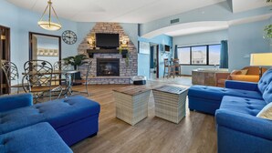 Una sala de estar con chimenea de piedra, sofás azules, mesas de centro de mimbre y una zona de comedor con sillas de metal.