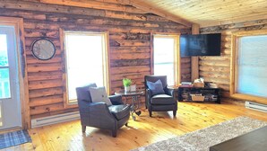 L’intérieur d’un chalet en rondins, avec deux fauteuils, une petite table sur laquelle est posée une plante, une horloge et un téléviseur accroché au mur.