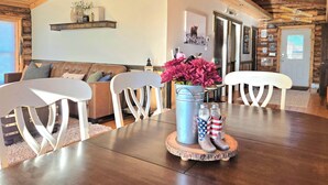Un salon chaleureux avec un plafond en bois, un canapé en cuir marron, des chaises blanches, une table basse avec un seau en métal et des bottes, et une porte ornée d’inscriptions.