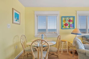 A dining area with a round glass table, wicker chairs, and a small stool. There are two windows with white blinds, a framed painting on the wall, and a lamp on the floor.