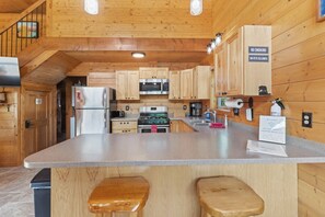 A wooden cabin interior with a kitchen area featuring a refrigerator, stove, microwave, and wooden cabinets. There are two stools at the counter, and a sign indicating no smoking and no pets allowed. - Kenai Cozy Cabin - 2 (Kenai)