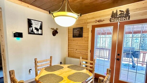 A rustic wooden dining area with a round table and chairs, a hanging light fixture, and a 'Welcome' sign above the door.