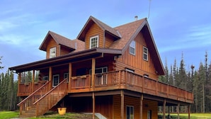 A large wooden cabin with multiple decks and a covered porch, surrounded by a grassy landscape and trees.