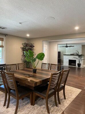 A dining area with a wooden table and chairs, a large potted plant, and a white fireplace in the background.