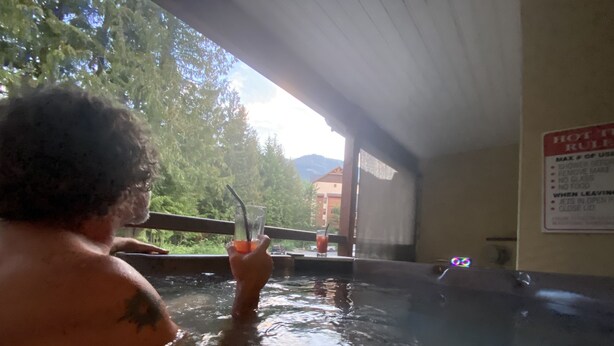 A person in a hot tub with a glass of drink, a sign with rules, and a view of trees and buildings.
