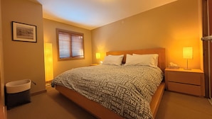 A neatly arranged bedroom with a large bed, bedside lamps, a bedside table, a waste basket, and a window with blinds.
