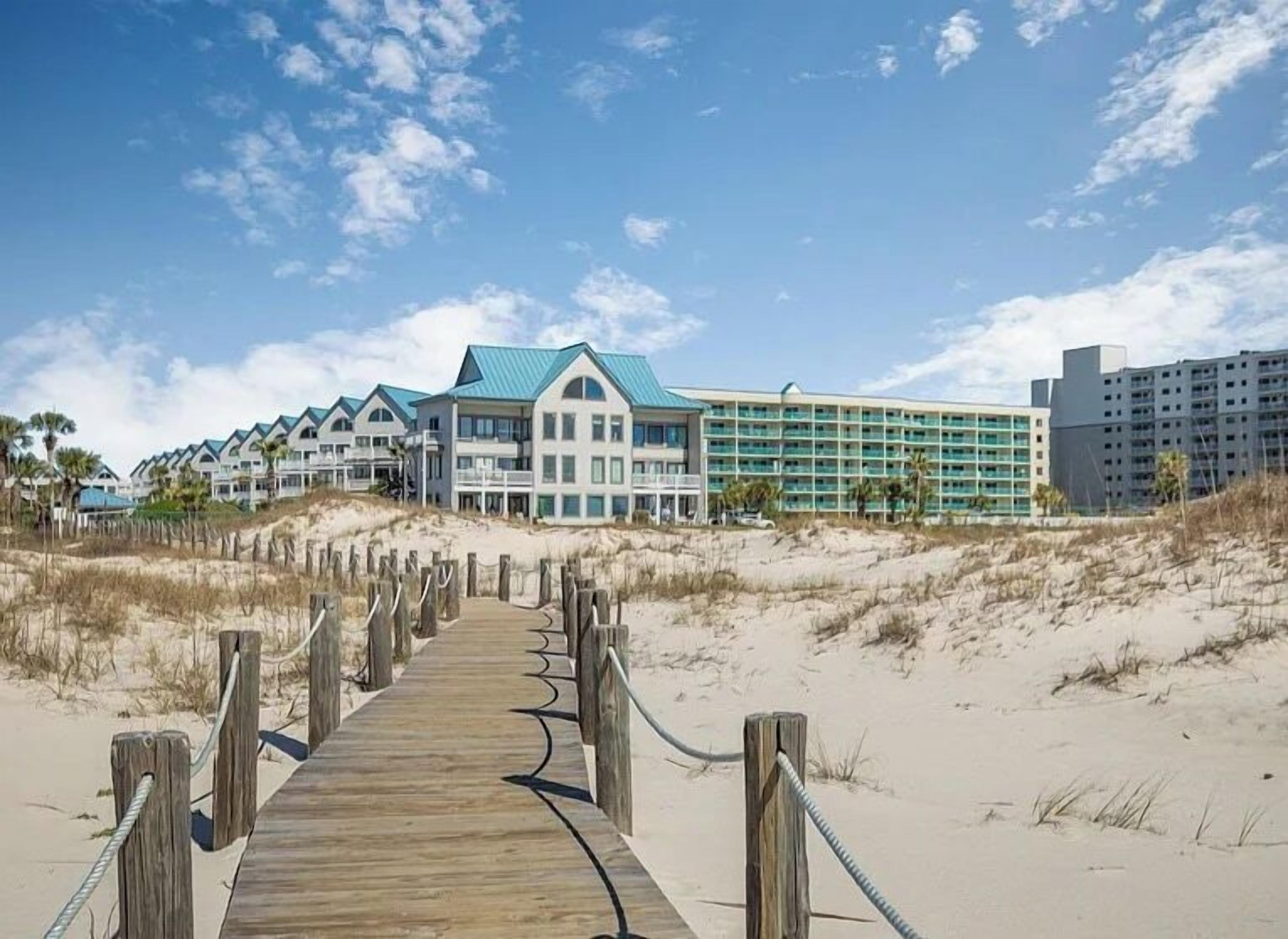 A beachside hotel with a wooden boardwalk leading to the entrance, featuring multiple buildings with blue roofs and balconies.
