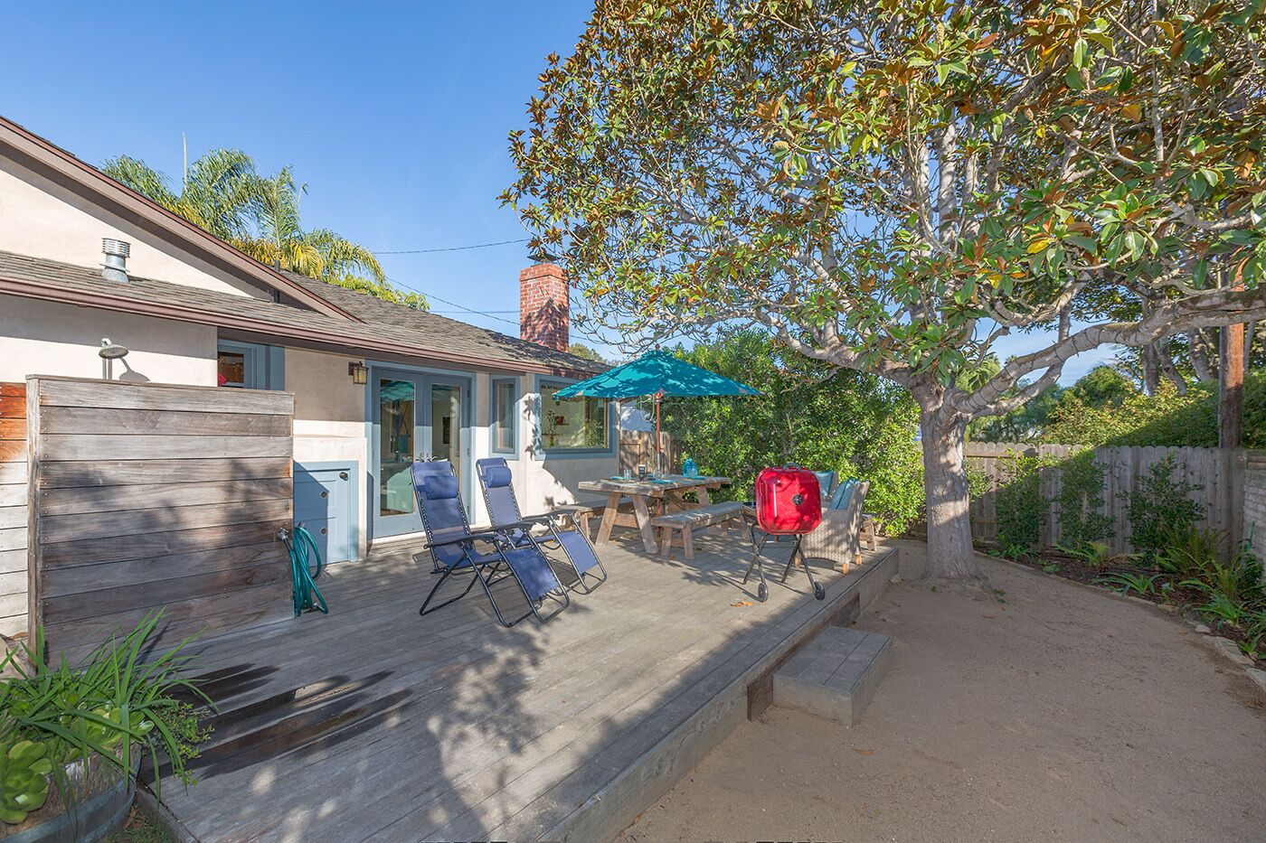 Un jardin avec une terrasse en bois, deux chaises longues, une table de pique-nique, un barbecue et un arbre qui offre de l’ombre.