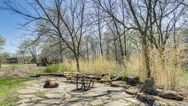 Un patio de piedra con mesas de picnic y un pozo de fuego, rodeado de árboles y rocas.