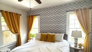 Ein Schlafzimmer mit einem Bett, einem Deckenventilator, einem Fenster mit Vorhängen, einer Nachttischlampe und einem kleinen Tisch mit Lampe.