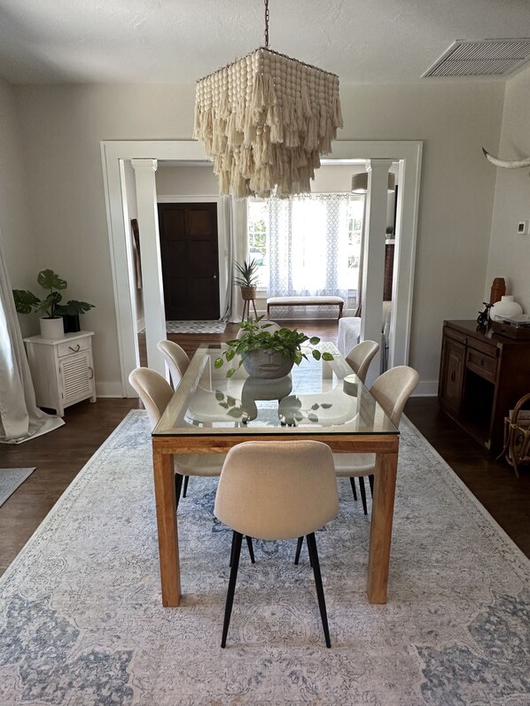 A dining area with a glass-top table, six chairs, a hanging light fixture, and a view of the outdoors through a large window.