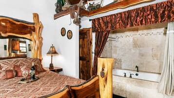 A bedroom with a wooden bed, a nightstand with a lamp, a large mirror, and a bathroom with a bathtub and tiled walls.